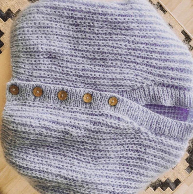 Pu&ograve; includere: Un cappello lavorato a maglia lilla chiaro con patta abbottonata. Il cappello &egrave; realizzato con un filato morbido e strutturato e presenta una fila di cinque bottoni in legno lungo la patta. Il motivo a maglia presenta strisce orizzontali.