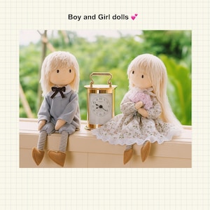 Puede incluir: Dos muñecos hechos a mano, un niño y una niña, están sentados uno al lado del otro. El muñeco del niño lleva un traje gris con una pajarita, mientras que la muñeca de la niña lleva un vestido floral. Un reloj dorado está entre ellos. El texto "Boy and Girl dolls" está arriba.