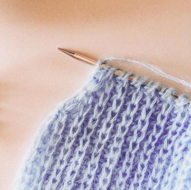 Pu&ograve; includere: Primo piano di lavoro a maglia in corso. Il progetto presenta filato blu chiaro su un ferro da maglia in legno. Il tessuto a maglia ha un aspetto strutturato, con punti visibili e una consistenza morbida e soffice. Lo sfondo &egrave; di colore neutro.