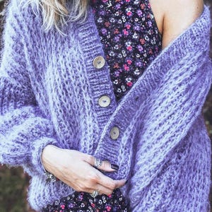 Pu&ograve; includere: Un cardigan lavorato a maglia grossa color lavanda con grandi bottoni in legno. Il design a trama aperta del maglione &egrave; visibile. Il cardigan &egrave; indossato sopra un vestito con stampa floreale.
