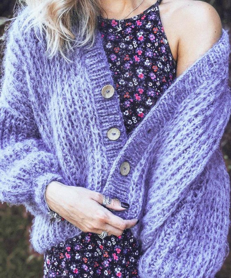 Pu&ograve; includere: Un cardigan lavorato a maglia grossa color lavanda con grandi bottoni in legno. Il design a trama aperta del maglione &egrave; visibile. Il cardigan &egrave; indossato sopra un vestito con stampa floreale.