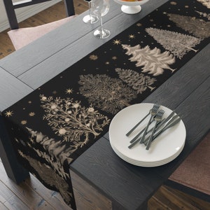 May include: A black table runner with a gold and brown design of stylised Christmas trees. The runner is on a dark wood table with a white plate and cutlery.