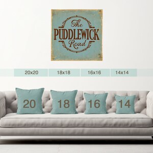 May include: A light blue and brown vintage-style sign with the text "The Puddlewick Road". The sign is displayed on a white wall with a sofa in the foreground. The sofa has four throw cushions with the numbers 20, 18, 16, and 14 printed on them.