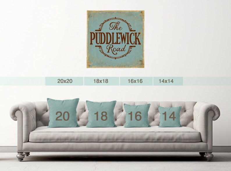 May include: A light blue and brown vintage-style sign with the text "The Puddlewick Road". The sign is displayed on a white wall with a sofa in the foreground. The sofa has four throw cushions with the numbers 20, 18, 16, and 14 printed on them.