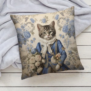 May include: A decorative cushion featuring a cat dressed in a blue and gold suit, holding a bouquet of white roses. The background is a cream-coloured floral pattern with blue flowers.