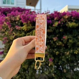 以下が含まれることがあります： オレンジと白の繰り返し花柄模様の布製ストラップが付いたリストレットキーホルダー。ストラップはゴールドトーンの金属製クランプで固定されており、鍵などを取り付けるための同色のゴールドトーンの円形スプリットリングが付いています。