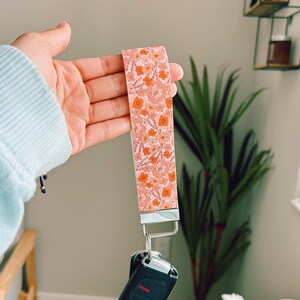 以下が含まれることがあります： ライトピンク、ピーチ、オレンジの色合いの花柄が繰り返される布製のリストレットキーホルダー。ストラップはシルバーカラーの金属製クラスプに取り付けられており、パニックボタンが付いた黒いプラスチック製の車のキーフォブがぶら下がっています。
