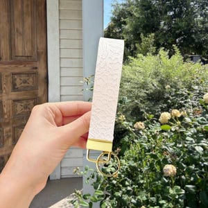 May include: A cream-colored wristlet key fob with a delicate lace pattern. The fob has a gold-colored metal accent and a matching key ring. The background includes greenery and a wooden door.