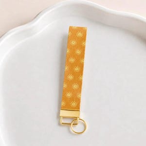 May include: A mustard yellow wristlet keychain strap featuring a repeating pattern of small, light yellow sun icons. The strap is attached to a polished gold-tone metal rectangular clamp and a matching circular split ring for holding keys.