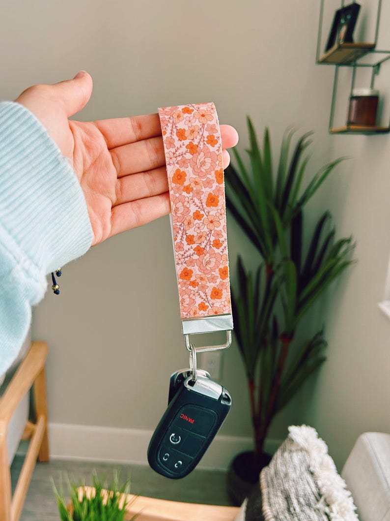 以下が含まれることがあります： ライトピンク、ピーチ、オレンジの色合いの花柄が繰り返される布製のリストレットキーホルダー。ストラップはシルバーカラーの金属製クラスプに取り付けられており、パニックボタンが付いた黒いプラスチック製の車のキーフォブがぶら下がっています。