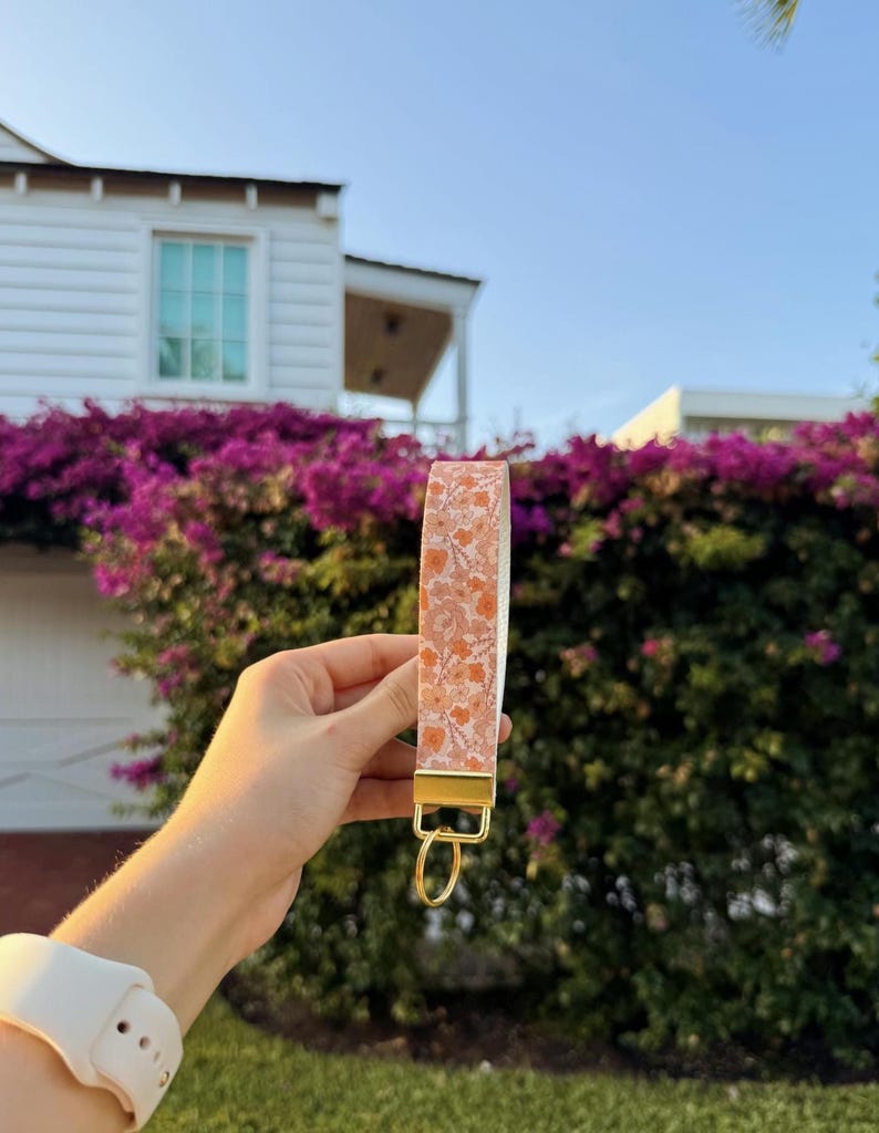 以下が含まれることがあります： オレンジと白の繰り返し花柄模様の布製ストラップが付いたリストレットキーホルダー。ストラップはゴールドトーンの金属製クランプで固定されており、鍵などを取り付けるための同色のゴールドトーンの円形スプリットリングが付いています。