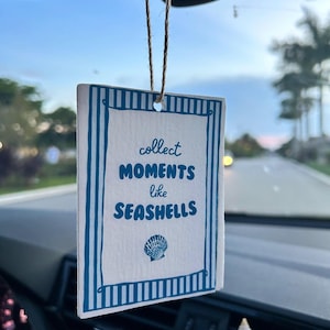 May include: A rectangular car air freshener hanging from a rearview mirror by a twine string. The white card features a blue striped border and the text "collect MOMENTS like SEASHELLS" in blue, with a small blue seashell illustration below.