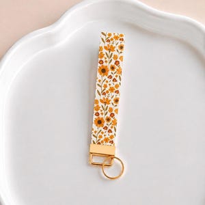 May include: A rectangular fabric wristlet keychain featuring a repeating pattern of orange, yellow, and brown flowers with green stems on a white background, finished with a gold-toned metal clamp and a matching circular split key ring.