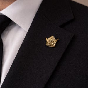 Könnte beinhalten: Ein goldfarbener Kronen-Ansteckpin auf einem schwarzen Anzug. Der Pin ist mit aufwendigen Designs versehen und befindet sich auf dem linken Revers, über einem weißen Hemdkragen. Nahaufnahme.