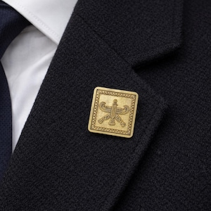 Könnte beinhalten: Ein goldfarbener quadratischer Ansteckpin mit einem eingravierten Design eines Vogels mit ausgebreiteten Flügeln, befestigt an einem dunkelblauen Blazer. Der Pin wird über einem weißen Hemd und einer marineblauen Krawatte getragen.