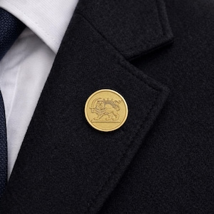 Op de afbeelding: Een goudkleurige ronde reversspeld met een gedetailleerd leeuw- en zon-embleem, bevestigd aan een donkergrijze wollen jas. De speld bevindt zich op de revers, in de buurt van een wit overhemd en een marineblauwe stropdas.