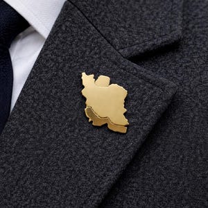 Könnte beinhalten: Ein goldfarbener Ansteckpin in Form des Iran, befestigt an einem dunkelgrauen Blazer. Der Pin zeigt die Umrisse des Landes. Der Blazer wird über einem weißen Hemd und einer marineblauen Krawatte getragen. Ein stilvolles Accessoire.