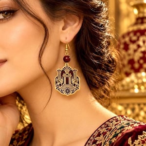 May include: A gold-toned dangle earring featuring a dark red bead and a flat, shield-shaped pendant. The pendant displays a detailed cutout design of two winged creatures flanking a central architectural structure with columns and a floral base.