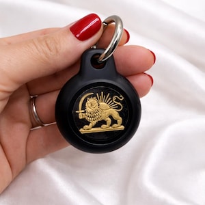 Puede incluir: Llavero redondo negro con un emblema dorado que representa un león sosteniendo una espada, con rayos de sol. El llavero tiene un anillo plateado. El diseño está centrado en la superficie negra.
