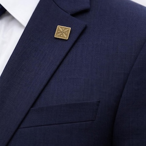 Puede incluir: Primer plano de una chaqueta de traje azul marino con un pin cuadrado dorado en la solapa. El pin tiene un diseño decorativo. Se ve una camisa blanca con cuello debajo.