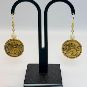 Puede incluir: Un par de pendientes dorados con diseño de moneda que presenta un león y un sol. Cada pendiente tiene una cuenta de color crema y un gancho dorado. Los pendientes se exhiben en un soporte negro sobre un fondo blanco.