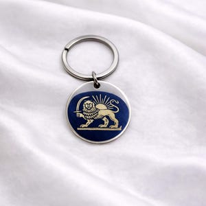 Puede incluir: Un llavero redondo con un anillo plateado y un emblema azul y dorado. El emblema presenta un león sosteniendo una espada, con un diseño de rayos de sol encima. El llavero está sobre un fondo blanco y sedoso.