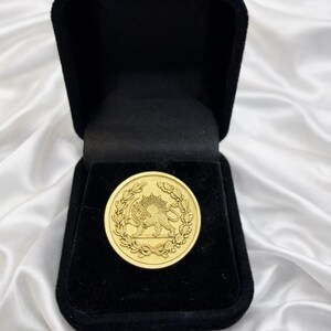 Puede incluir: Una moneda de oro con un emblema de león y sol, presentada en una caja de terciopelo negro abierta. La moneda está rodeada por un diseño de corona de laurel. El fondo es una tela blanca y sedosa.