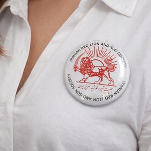 May include: A white button pin with the words "IRANIAN RED LION AND SUN SOCIETY" in a circular design. The pin features a red lion holding a sword and a rising sun. The pin is pinned on a white shirt.
