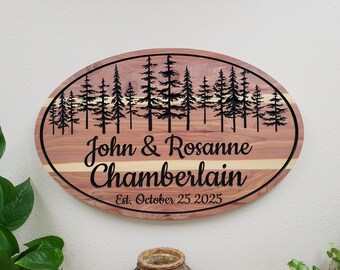 May include: An oval wooden sign featuring a laser-engraved forest of pine trees across the top. The text reads John & Rosanne Chamberlain and Est. October 25 2025 in black script and serif lettering. The wood has a natural reddish-brown grain.