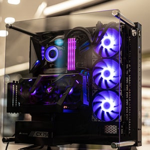 Puede incluir: Una caja de ordenador negra con un panel lateral de vidrio, que muestra un sistema de iluminación RGB morado y azul. La caja tiene una fuente de alimentación Corsair CX750.