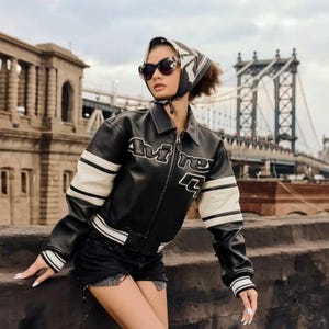 May include: A black leather jacket with white stripes on the sleeves and the text Avi Rex on the chest. The jacket features a collar, a front zipper, and ribbed cuffs. The garment is paired with a patterned headscarf and dark sunglasses.