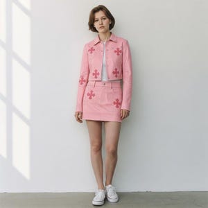 May include: A light pink cropped jacket and matching mini skirt set featuring dark pink cross-shaped appliqués. The jacket has a collared neckline, long sleeves, and silver-toned button closures. The skirt features a straight hem and button closure.