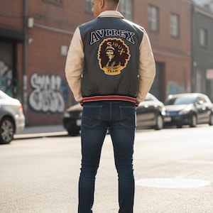 Peut inclure: Une veste universitaire avec un corps en laine grise et des manches en cuir crème. Le dos présente un écusson brodé avec le texte AVIREX SIOUX FOOTBALL TEAM et un graphique représentant une personne. La veste a un col et des poignets côtelés.