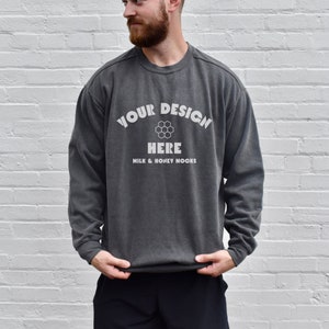 Könnte beinhalten: Ein anthrazitfarbener Sweatshirt mit langen Ärmeln und einem Rundhalsausschnitt. Der Sweatshirt hat ein weißes Grafikdesign mit dem Text "YOUR DESIGN HERE" mit einem Wabenmuster und dem Text "MILK & HONEY NOOKS" darunter.