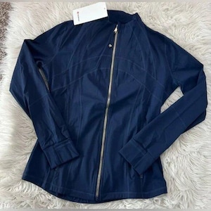May include: A navy blue jacket with a full-length gold zipper and a stand-up collar. The jacket has long sleeves and a fitted silhouette. The seams are subtly stitched, and a small tag is attached near the collar.