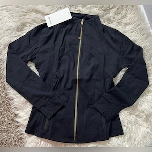May include: Black zip-up jacket with a high collar and gold-colored zipper. The jacket has a fitted silhouette and long sleeves. A white tag is attached to the collar. The jacket is displayed on a white and tan textured surface.