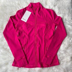 May include: A vibrant pink athletic jacket with a full-length zipper, long sleeves, and a high collar. The jacket has a fitted design with subtle stitching details and a small tag attached. The jacket is made of a smooth, lightweight material.