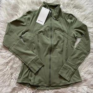 May include: Olive green zip-up jacket with a high collar and thumbholes. The jacket has a fitted design with seams and a tag attached. The jacket is made of a soft, stretchy material. The jacket is a sporty style.
