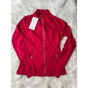 May include: A vibrant red zip-up jacket with a high collar and gold-colored zipper. The jacket has a fitted design with seams and a tag attached. The jacket is displayed on a white, fluffy surface.