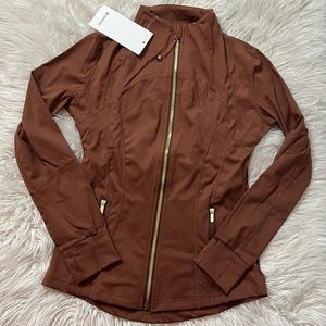 Può includere: Giacca marrone con collo alto e cerniera dorata a tutta lunghezza. La giacca ha due tasche con cerniera e un'etichetta attaccata al colletto. La giacca è realizzata in un materiale liscio e leggero.