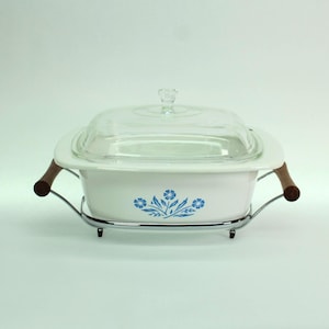 May include: A white rectangular ceramic casserole dish with a clear glass lid. The dish features a blue floral pattern on the front and is set in a chrome metal cradle with two brown wooden handles on the sides for easy lifting and serving.