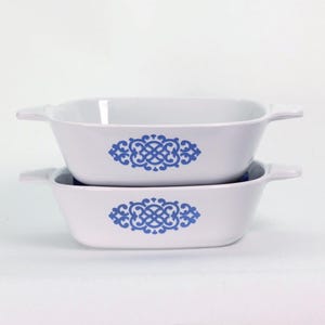 May include: Two white rectangular baking dishes with handles are stacked. Each dish features a blue decorative design in the center. The dishes are made of ceramic and are suitable for oven use.