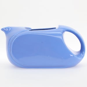 May include: A light blue ceramic pitcher with a rounded, modern design. The pitcher has a wide, flat base and a spout on one side. It has a handle on the opposite side.