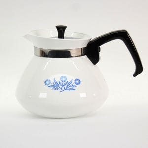 May include: A white teapot with a wide, rounded base and a tapered neck. It features a blue floral pattern on the front, a black handle, a black lid knob, and a silver-colored metal band encircling the upper section of the teapot body.