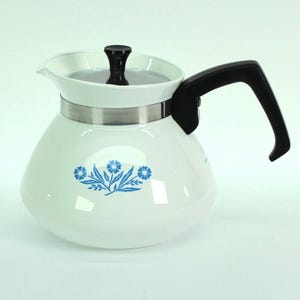 May include: A white teapot with a rounded body and a blue floral pattern on the front. It features a black handle, a black lid knob, and a metallic silver band around the upper neck. The design includes a small spout for pouring liquids.