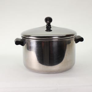Op de afbeelding: Een roestvrijstalen kookpot met een deksel en twee handvatten aan de zijkant. Het deksel heeft een zwarte knop bovenop. De pot staat op een wit oppervlak. De pot is ontworpen om te koken.