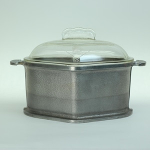 May include: A vintage, square-shaped casserole dish with a clear glass lid. The dish is made of a textured, gray metal and has a handle on each side. The lid has a decorative handle and a diamond-shaped design.