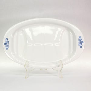 May include: An oval white ceramic serving platter featuring a smooth, glossy finish and two recessed sections on the left and right sides. The platter is decorated with a blue floral pattern on both ends, consisting of small flowers and leaves.