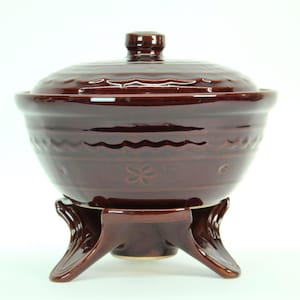 May include: A brown ceramic fondue pot with a lid and a stand. The pot has a wavy design around the middle and a floral pattern. The stand has three legs.
