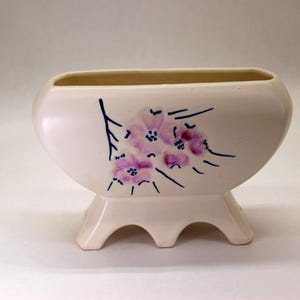 May include: A white ceramic planter with a floral design. The planter has a rectangular shape with a rounded bottom and three legs. The flowers are pink and blue.